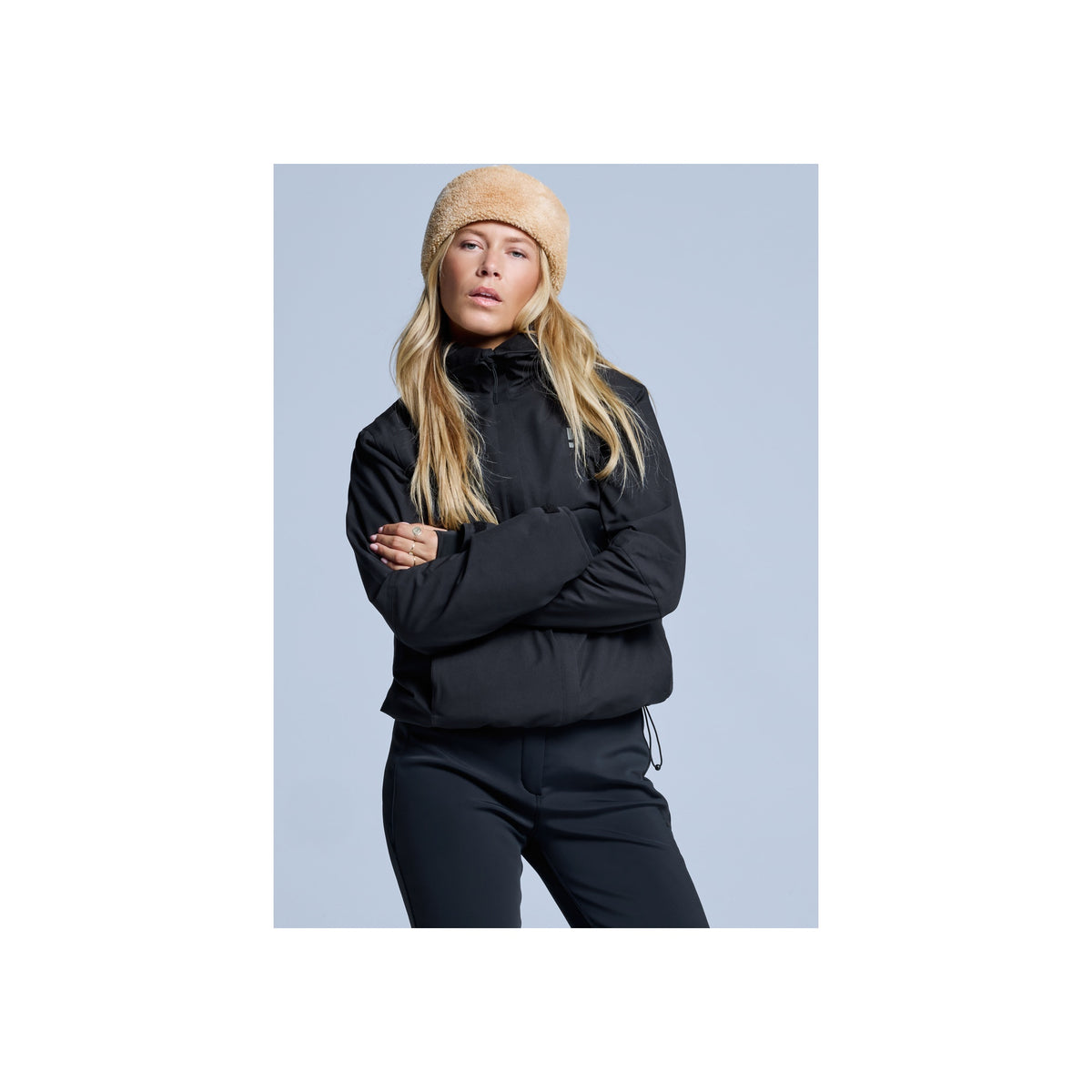 Blonde vrouw draagt zwarte halfzip sweater en beige muts, armen over elkaar, casual sportieve look