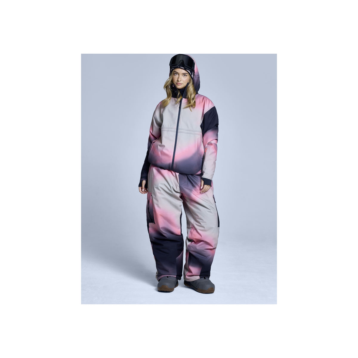 Vrouw in roze-grijs-zwart ski-onesie met capuchon en winterlaarzen op lichte achtergrond