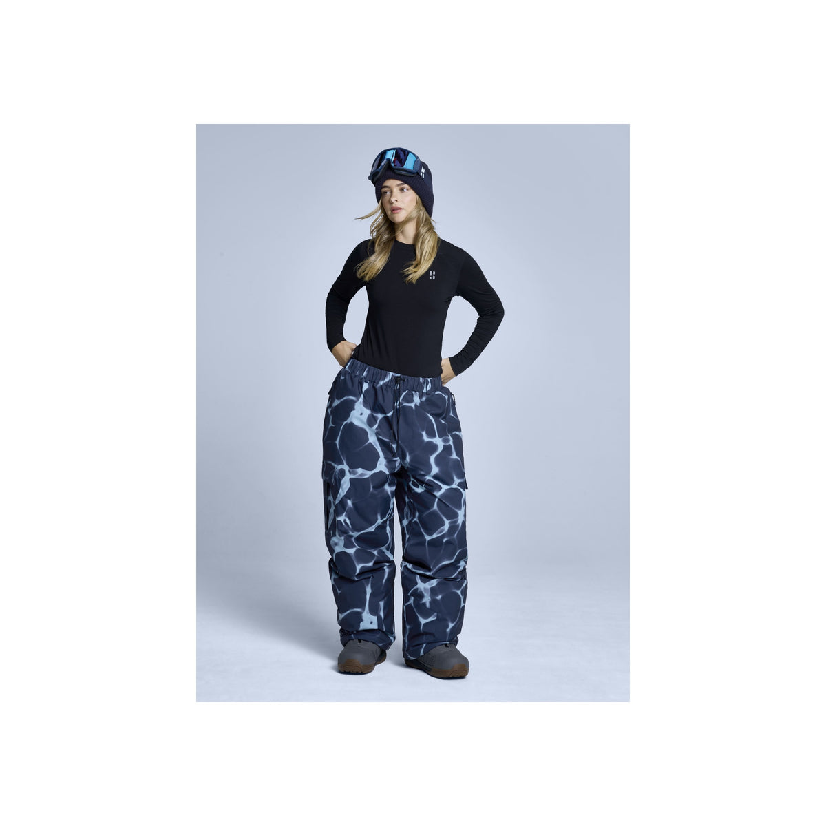Vrouw in zwarte trui en blauwe camouflage skibroek met bijpassende muts, volledige lengte studiofoto